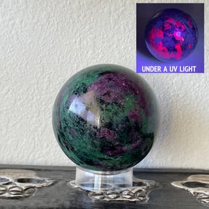 May include: Green and pink gemstone sphere on a clear stand. The sphere glows pink under a UV light. "UNDER A UV LIGHT" is written below the sphere.