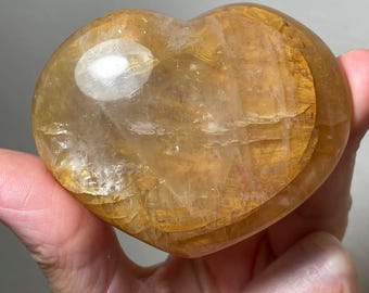 May include: A heart-shaped, polished citrine crystal with a golden yellow hue. The crystal has a translucent appearance with visible internal striations.