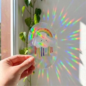 Op de afbeelding: Een heldere, ovale raamsticker met een regenboog en een lachende wolk met kleurrijke regen. De sticker creëert een regenboogeffect wanneer er licht doorheen schijnt. De achtergrond omvat een raam, een plant en een witte muur.