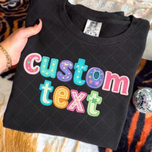 以下が含まれることがあります： キルティング模様の黒いスウェットシャツ。カラフルな刺繍文字で「CUSTOM TEXT」と書かれています。文字は様々な色で縁取られ、星と月のアクセントが付いています。