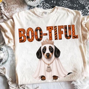 Może przedstawiać: Kremowy t-shirt z pomarańczowo-czarnym napisem BOO-TIFUL. Pod tekstem znajduje się grafika jamnika ubranego w biały kostium ducha, złotą koronę oraz złoty naszyjnik z małą zawieszką w kształcie ducha zwisającą z łańcuszka.