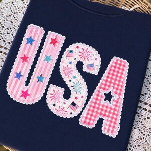 May include: A navy blue t-shirt displaying the word "USA" in a decorative appliqu&eacute;. The letters are adorned with pink stripes, fireworks, and a red and white gingham pattern, with star accents and a scalloped edge.