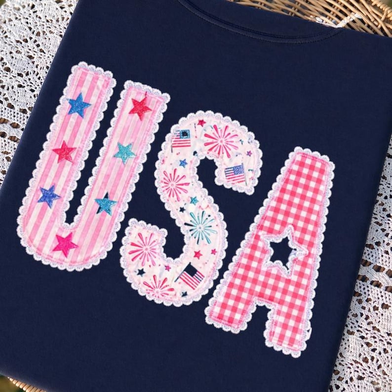 May include: A navy blue t-shirt displaying the word "USA" in a decorative appliqu&eacute;. The letters are adorned with pink stripes, fireworks, and a red and white gingham pattern, with star accents and a scalloped edge.