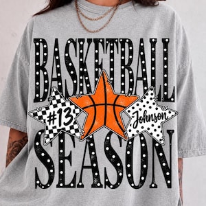 以下が含まれることがあります： 白い水玉模様の大きな黒い文字で「BASKETBALL SEASON」と書かれたグレーのTシャツ。文字の間には3つの星があり、#13と書かれたチェック柄の星、バスケットボール柄のオレンジ色の星、Johnsonと書かれた白い星が配置されています。