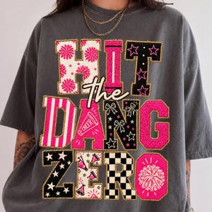 以下が含まれることがあります： 「HIT The DANG ZERO」という文字が大きくプリントされたチャコールグレーのTシャツ。文字はピンクと黒のパターンで装飾されており、ポンポン、星、リボン、ストライプ、市松模様などが描かれ、ゴールドの縁取りが施されています。