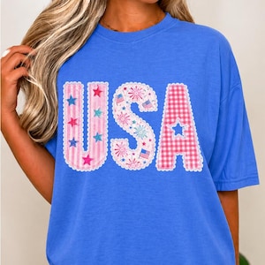 May include: A blue t-shirt featuring the word "USA" in a patriotic design. The letters are decorated with stars, fireworks, and a gingham pattern, ideal for celebrating Independence Day.