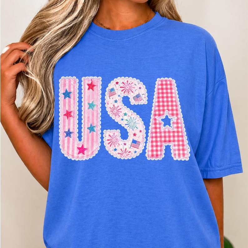 May include: A blue t-shirt featuring the word "USA" in a patriotic design. The letters are decorated with stars, fireworks, and a gingham pattern, ideal for celebrating Independence Day.