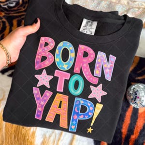 Puede incluir: Sudadera negra con la frase "BORN TO YAP!" en letras coloridas y estrellas. La sudadera tiene un diseño acolchado y cuello redondo. Una mano con una pulsera dorada sostiene la sudadera.