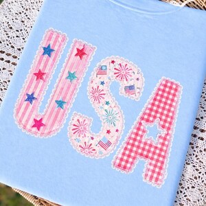 May include: Pale blue t-shirt featuring the word "USA" in appliqued letters. The letters are adorned with patriotic designs: pink striped fabric and stars for the "U", fireworks and flags for the "S", and red gingham with a star cut-out for the "A".