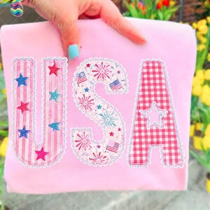 May include: A pale pink t-shirt featuring the letters "USA" in a patriotic design. The letters are adorned with stars, fireworks, and American flags. The "U" has pink stripes, the "S" has fireworks, and the "A" has a red and white gingham pattern.
