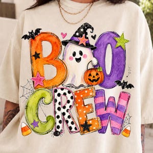 May include: A cream-colored t-shirt featuring a colorful graphic with the text BOO CREW. The design includes a white ghost wearing a black witch hat, a pumpkin bucket, bats, spiderwebs, stars, and candy corn in orange, purple, and green hues.