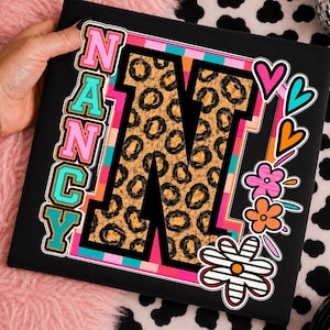 May include: A black t-shirt featuring a large letter N with a brown and black leopard print pattern. The name Nancy is written vertically in pink and turquoise block letters. The design includes colorful hearts and flowers on the right side.