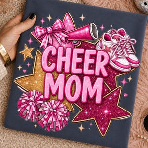Könnte beinhalten: Ein dunkelgrauer Stoff mit einem Grafikdruck, der den Text CHEER MOM in Pink zeigt. Das Design umfasst ein pinkes Megafon, zwei pinke Pompons, ein Paar pink-weiße Sneaker sowie goldene und pinke glitzernde Sterne mit kleinen Funken.
