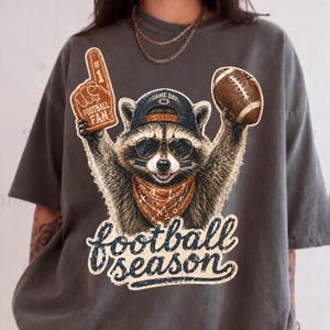 Op de afbeelding: Een antracietgrijs t-shirt met een afbeelding van een wasbeer met een baseballpet en zonnebril, die een nummer één schuimvinger en een football vasthoudt. De wasbeer draagt een bandana en de tekst eronder luidt football season in cursief schrift.