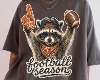 May include: A charcoal gray t-shirt featuring a graphic of a raccoon wearing a baseball cap and sunglasses, holding a number one foam finger and a football. The raccoon wears a bandana, and the text below reads football season in script.