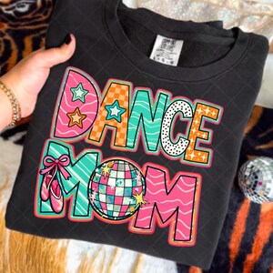 以下が含まれることがあります： カラフルなブロックレターで「DANCE MOM」と書かれた黒いスウェットシャツ。デザインには星、ディスコボール、バレエシューズが含まれています。文字はピンク、オレンジ、ティール、白です。