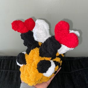 Könnte beinhalten: Ein handgefertigter, plüschiger Hut im Hummel-Design. Der Hut hat einen gelben Körper mit schwarzen Punkten, weißen Akzenten und roten herzförmigen Antennen. Der Hut ist aus weichem, strukturiertem Garn gefertigt.