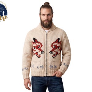 May include: A cream-coloured zip-up cardigan with red and black fox designs on the front. The sweater features a shawl collar, pockets, and blue and white accents on the sleeves. The model is wearing blue jeans.