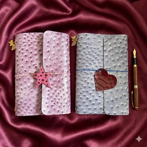 May include: Two rectangular journals with textured, ostrich-like covers in light purple and light blue. The purple journal features a pink star charm, while the blue one has a dark red heart charm. Both include small gold butterfly charms and a fountain pen.