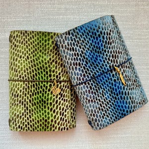 Op de afbeelding: Twee rechthoekige notitieboeken met een gestructureerd stippenpatroon. Het linker notitieboek is groen met een gouden zwaan-bedel aan een donker elastiek. Het rechter notitieboek is blauw met een gouden veer-bedel aan een donker elastiek. Ovaal patroon.