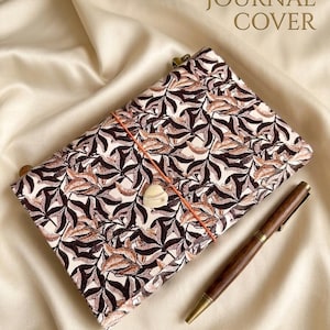 Op de afbeelding: Een lederen journal cover met een crèmekleurige achtergrond en een dicht patroon van donkerbruine en beige bladeren. Een oranje elastisch koord zit om de cover, vastgezet met een kleine, lichtgekleurde hartvormige bedel. Een donkerhouten pen ligt ernaast.