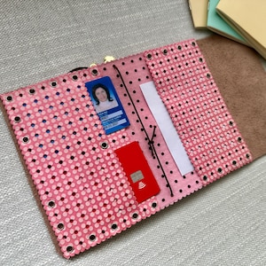 May include: A pink leather wallet featuring a perforated floral pattern with silver metal eyelets along the edges. The interior includes a blue identification card, a red credit card, and a white paper insert held by a thin black elastic cord.