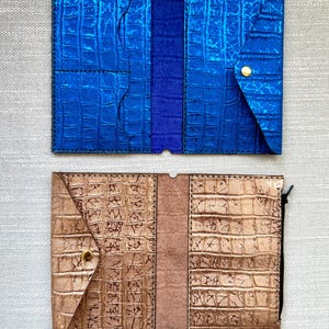 May include: Two open wallets with a textured crocodile-skin pattern. The top wallet is metallic blue with a gold-tone snap closure and interior card slots. The bottom wallet is metallic bronze with a gold-tone snap closure and a black side zipper.