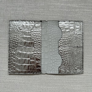 May include: A metallic silver card holder featuring a textured crocodile skin pattern on the outer panels and a smooth, light gray central section. The accessory includes two vertical slots on each side with curved cutouts for easy card access.
