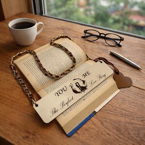 Puede incluir: Un libro abierto con una cadena decorativa y un marcapáginas que dice "YOU & ME Love Story The Perfect". Una taza de café, gafas y un bolígrafo están sobre una superficie de madera.