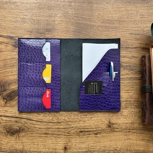 May include: A purple leather notebook cover with a textured finish, featuring three card slots on the left side and a pocket for a notepad, a pen holder, and a small black card slot on the right side, with three cards and a pen inserted.