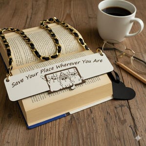 Puede incluir: Un libro abierto con un asa de cadena dorada y negra. Un marcapáginas blanco con el texto "Save Your Place Wherever You Are" y una ilustración de gato descansa sobre las páginas. Una taza de café, gafas y un lápiz están sobre una superficie de madera.