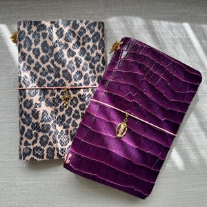 Può includere: Due quaderni rettangolari con chiusure elastiche. Uno presenta un motivo leopardato beige e nero con un ciondolo a fiocco dorato. L'altro è viola intenso con una trama a pelle di coccodrillo e un ciondolo ovale dorato con una figura.