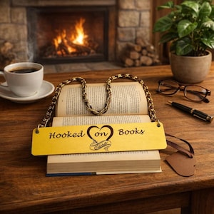 Op de afbeelding: Een gele houten bladwijzer met de tekst "Hooked on Books" en een hartontwerp, rustend op een open boek. De bladwijzer heeft een gouden ketting en bruine lederen accenten. Een kopje koffie en een open haard zijn op de achtergrond.