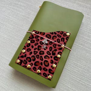 Puede incluir: Una funda para cuaderno de color verde oliva con un bolsillo rectangular de estampado de leopardo rojo y negro. El bolsillo está sujeto con remaches metálicos dorados y una banda elástica rosa claro que sostiene un dije plateado de bicicleta.