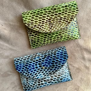 Pode incluir: Duas pequenas carteiras de couro feitas à mão. Uma é verde com manchas marrons escuras, a outra é azul com manchas pretas. Ambas têm um fecho de aba com um pequeno botão de pressão dourado.