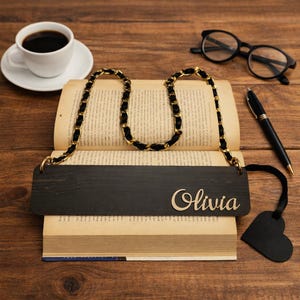 Puede incluir: Un marcapáginas de madera negra con el nombre "Olivia" en letras doradas, conectado a una cadena negra con detalles dorados. El marcapáginas está en un libro abierto, con una etiqueta negra en forma de corazón.