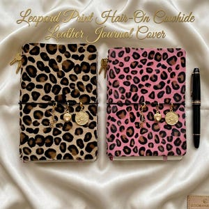 May include: Two rectangular journal covers made of hair-on cowhide leather with a leopard print pattern. One cover is tan and black, the other is pink and black. Each features a black elastic band with three gold-tone charms and a black pen.