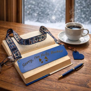 Puede incluir: Un marcapáginas de cuero azul con la frase "Sorry, I'm Fully Booked" en un libro abierto. Una cinta estampada, una taza de café, un bolígrafo y un dije en forma de corazón están sobre una mesa de madera.