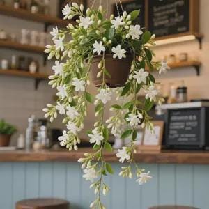 May include: A hanging brown pot filled with white flowers and green leaves. The cascading plant is suspended from a hook, with the flowers and foliage spilling downwards. The background includes a wooden shelf and a counter.