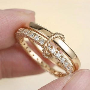 May include: A gold-colored ring with a knot design. One band is encrusted with small, clear gemstones. The ring has a polished finish and is held by a person's fingers. The brand name "YUVANA JEWELS" is visible.