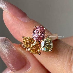 Puede incluir: Un anillo de oro con un grupo de piedras preciosas de colores. El anillo presenta una disposición de piedras preciosas rosas, amarillas y verdes. El anillo está engastado con un óvalo rosa, una marquesa amarilla y una piedra verde en forma de pera. El anillo está etiquetado como "YUVANA JEWELS".