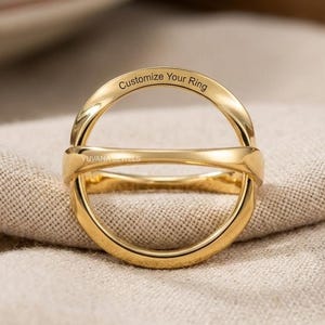 May include: A gold-colored ring with a unique, open design. The ring features the text "Customize Your Ring" and the brand name "YUVANA JEWELS". The ring is displayed on a beige fabric surface.