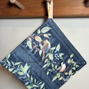 May include: A square quilted potholder featuring a dark blue fabric background with a printed pattern of birds, green leaves, and dark berries. The potholder is stitched with a diamond pattern and includes a fabric loop for hanging.