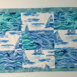 May include: A rectangular quilted placemat featuring a nautical theme with alternating blocks of blue and teal ocean waves and white fabric panels printed with sailboats. The entire piece is finished with a solid light blue fabric border.