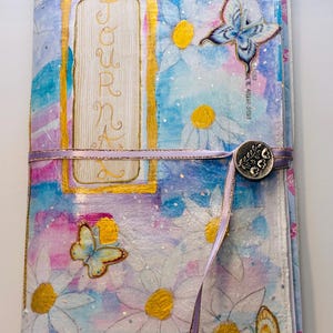 May include: Handmade journal with a watercolor design featuring daisies and butterflies. The cover has a gold-framed rectangle with the word "Journal" in gold script. A lavender ribbon wraps around the journal, secured with a decorative button.