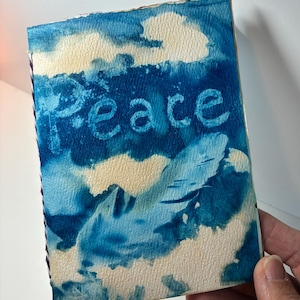 May include: A handmade greeting card with the word "Peace" in blue lettering. The card features a watercolor design with blue and off-white hues, resembling clouds. The edges are slightly rough, giving it a rustic feel.
