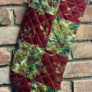 May include: A quilted Christmas stocking featuring a patchwork design of alternating squares. One set of squares displays a dark red fabric, while the other set features a green pattern of pine needles, pinecones, and holly leaves.