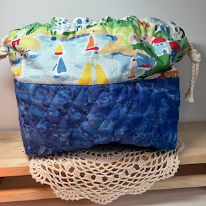 May include: A drawstring pouch featuring a top section with a colorful coastal landscape print of sailboats and buildings, and a bottom section made of quilted dark blue fabric. The bag is cinched with thick, cream-colored rope drawstrings.