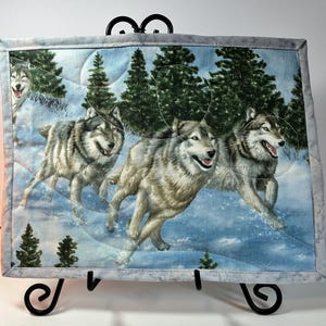 May include: A rectangular fabric wall hanging featuring a printed scene of four grey wolves running through a snowy forest with evergreen trees. The quilted piece has a light grey border and is displayed on a black metal stand.