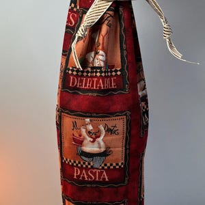 May include: Red fabric wine bottle bag with a pasta theme. The bag features illustrations of chefs, pasta dishes, and the words "Delectable" and "Pasta". A gold and white ribbon is tied at the top.